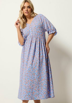 Blondynka w niebieskiej sukience midi w różowy kwiatowy wzór, z guzikami na gorsecie, bufkami i marszczoną talią, pozująca na białym tle.