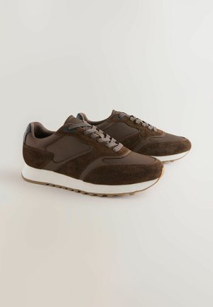 Pruunid tossud seemisnahast ja tekstiilist pealses, tekstuursete detailide, paelte ja valge vaherõhuga. Omavad kummist välistallaga haarde tagamiseks.