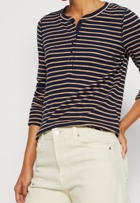 Granatowo-beżowa koszula henley w paski z długim rękawem, zapinana na guziki, z prążkowaną fakturą, zestawiona z jasnymi spodniami.