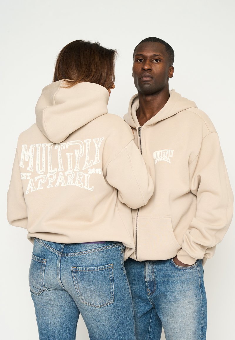 Sudaderas con capucha beige con un bolsillo canguro, con el texto blanco "MULTIPLY APPAREL" impreso en la parte trasera y en el pecho. Combinadas con jeans azules.