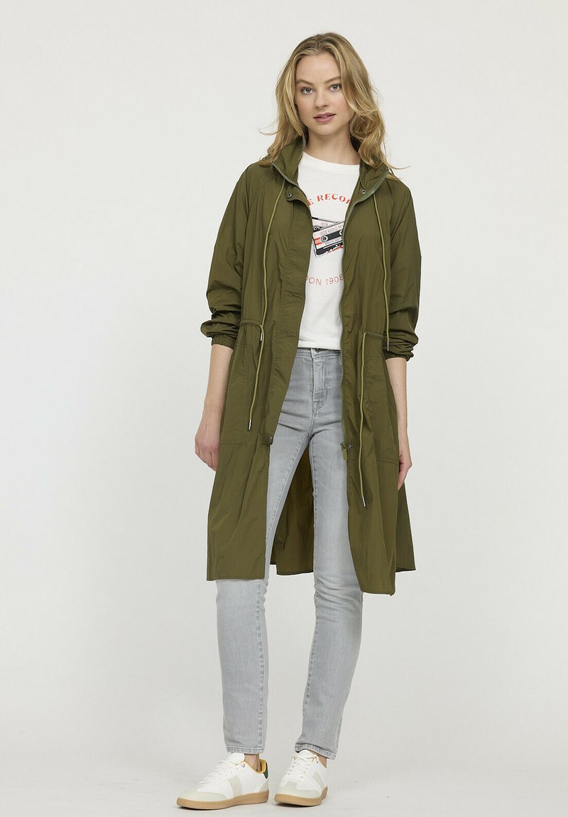 Veste légère olive avec une taille cintrée, fermeture éclair à l'avant et capuche, associée à un jean gris et des baskets blanches.
