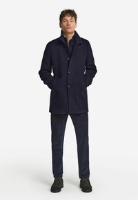 Marineblauer Wollmantel mit hohem Kragen, Knopfverschluss und Seitentaschen; getragen mit dunklen Kordhosen und schwarzen Schuhen.