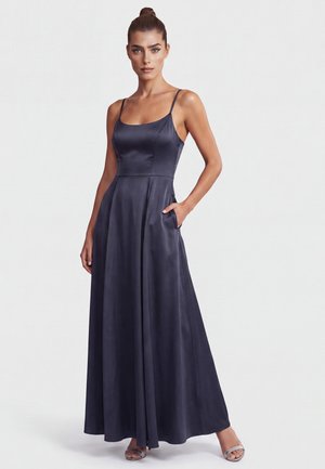 Woman with hair in a bun wearing a sleeveless dark navy satin maxi dress with thin straps and sandals, standing with one hand in pocket.