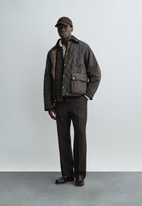 Moški, oblečen v temno prešito jakno z velikim žepom, rjave hlače, rjavi čevlji in rjava kapa s šiltom, stoji na sivi talni površini.