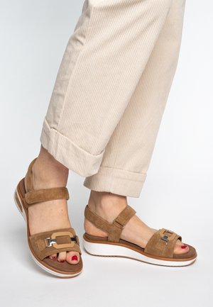 Feet wearing beige suede sandals with velcro straps and metallic buckles, paired with cream-colored cuffed corduroy pants on a white background.