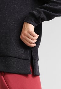 Sweat-shirt noir tacheté avec une fermeture éclair latérale. Comprend des poignets côtelés et un patch logo. Tissu texturé et coupe décontractée.