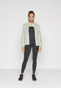 Ανοιχτό γκρι τζάκετ με φερμουάρ μπροστά, με ψηλό γιακά, ελαστικούς καρπούς και απαλή υφή, φορεμένο πάνω από μαύρο τοπ και μαύρα κολάν.