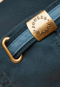 Gros plan sur une sangle en tissu bleu avec une boucle en laiton gravée "Joules" et une boucle en laiton sur un matériau texturé bleu foncé.