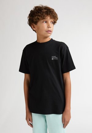 Camiseta negra de algodón con mangas cortas, cuello redondo y un pequeño diseño blanco impreso en el pecho. Llevada con pantalones azul claro.