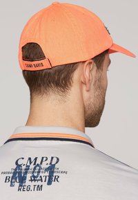 Cappellino da baseball arancione brillante con una fascia in tessuto, con il testo "CAMP DAVID" sul retro. Maglietta grigia con accenti blu e arancioni.