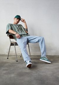 Chemise surdimensionnée rayée verte, jeans bleu clair, baskets vertes avec des accents blancs, assis sur une chaise en métal contre un arrière-plan neutre.