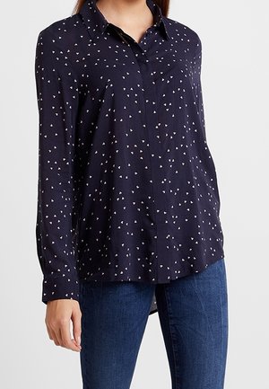 Donna che indossa una camicia blu navy con bottoni e piccoli motivi a cuore bianchi e jeans blu, in piedi contro uno sfondo chiaro e semplice.