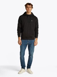 Giovane uomo in piedi, che indossa un felpa nera con tasca frontale, jeans blu e sneakers bianche, su uno sfondo bianco semplice.