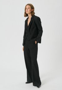 Black pinstriped suit featuring a fitted blazer, notch lapels, and wide-leg trousers. Material has a smooth texture. Black patent shoes complete the look.