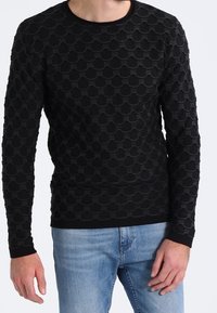 Pull noir à manches longues avec un motif texturé en vagues. Poignets et encolure côtelés, associé à un jean bleu clair.