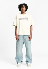 Kremfarget oversized t-skjorte med teksten "Reternity", sammen med lyseblå baggy jeans og hvite sneakers, står mot en ensfarget bakgrunn.
