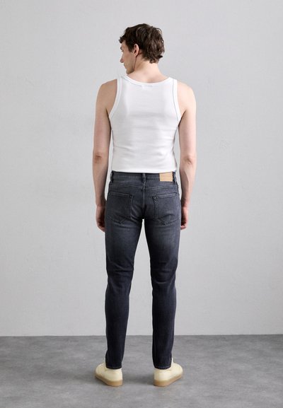 Jeans de mezclilla oscuros, de corte ajustado, con bolsillos traseros, combinados con una camiseta sin mangas blanca y zapatillas amarillas claras. Diseño minimalista, sin patrones visibles.