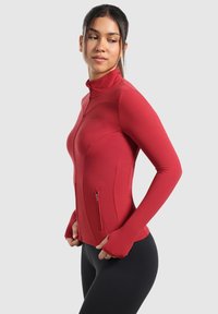 Rød sporty zip-up jakke lavet af strækbart materiale, med høj krave, tommelhulærmer og en lille lynlåslomme foran.