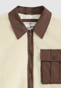 Chaqueta Sherpa con un cuerpo color crema, cuello marrón y cierre de cremallera. Cuenta con un bolsillo frontal con solapa, texturas contrastantes y etiqueta de la marca.
