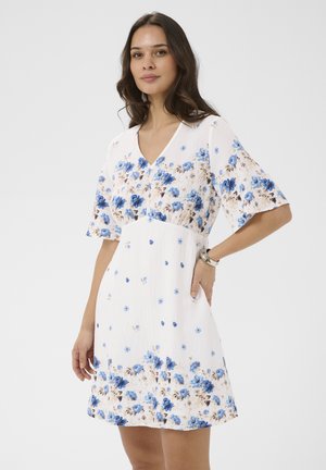 Žena v bielych šatách po kolená s modrým kvetinovým vzorom, s výstrihom do V a rukávmi po lakte, pózujúca s jednou rukou v vrecku.