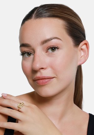 Frau mit heller Haut und zurückgebundenen braunen Haaren, trägt einen goldfarbenen, strukturierten quadratischen Ring an ihrer linken Hand nahe der Schulter, neutraler Hintergrund.