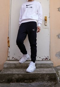 Person står på betongtrappor iförd vit Nike Air-tröja, svarta Nike Air-joggingbyxor och vita Nike-sneakers med snörning.