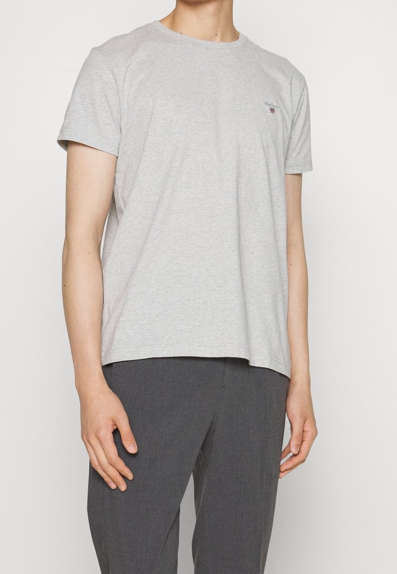Homme portant un T-shirt à manches courtes gris clair avec un petit logo sur la poitrine et un pantalon gris foncé, debout devant un fond blanc.
