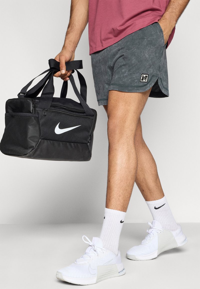 Schwarze Nike-Reisetasche mit weißem Logo, graue Shorts mit einem Logo-Patch, weiße Turnschuhe und weiße Socken mit schwarzem Logo.