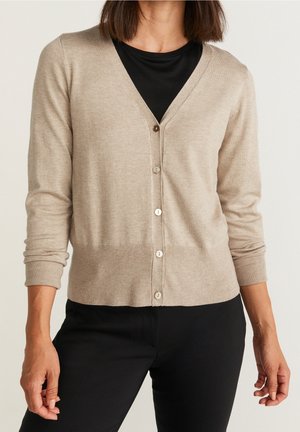 Kobieta ubrana w beżowy sweter na guziki nałożony na czarną bluzkę i czarne spodnie, stojąca na jednolitym tle.