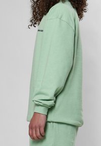 Felpa verde menta con maniche lunghe, polsini a coste e vestibilità rilassata. Presenta del testo nero sul davanti. Tessuto morbido e design semplice.