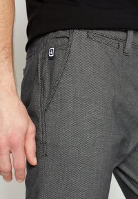 Gros plan sur la main d'une personne reposant près de la poche d’un pantalon gris texturé avec une petite étiquette bleu marine montrant une flèche blanche vers le bas.