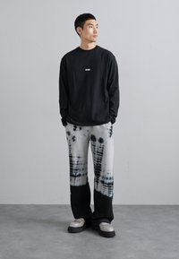 Schwarzes langärmliges Shirt mit kleinem weißen Logo, kombiniert mit grauen Batik-Hose im weiten Beinstil mit schwarzen Akzenten und entspannter Silhouette.