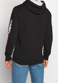 Zwarte hoodie van katoen met een kangoeroezak; beschikt over witte "TOMMY" tekst op de mouw en een grijze binnenvoering in de capuchon.
