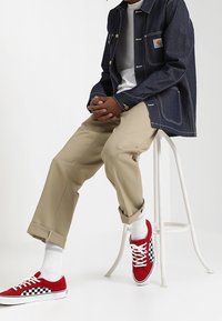 Osoba v tmavé džínové bundě, bílém tričku, béžových kalhotách, bílých ponožkách a červených teniskách s černobílými kostkovanými stranami, sedí na bílé stoličce.