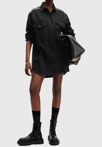 Personne portant une robe-chemise noire surdimensionnée à boutons avec poches poitrine, bottes de combat noires, chaussettes noires transparentes, tenant une grande pochette noire.