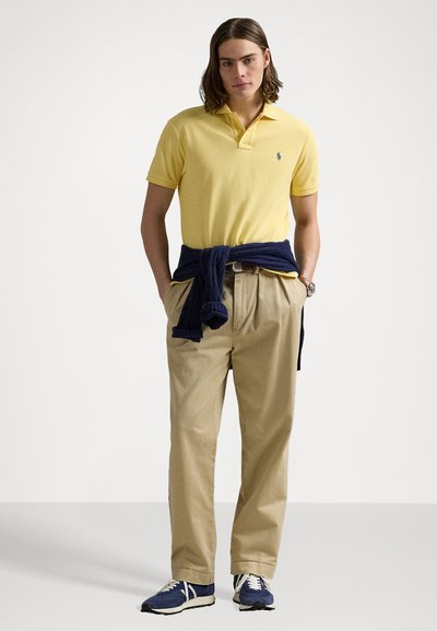 Cămașă polo galbenă cu un logo mic, asociată cu pantaloni bej. Pulover bleumarin legat la talie și adidași albaștri cu accente albe.