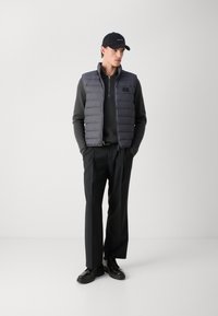 Grau gepolsterte Weste mit einem Reißverschluss auf der Vorderseite, schwarzes Langarmshirt, schwarze Hose und schwarze Schuhe. Enthält eine schwarze Kappe mit einem Logo.