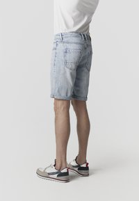 Homme portant un short en denim bleu clair, un t-shirt blanc et des baskets blanches avec des accents noirs et rouges, debout devant un fond uni.