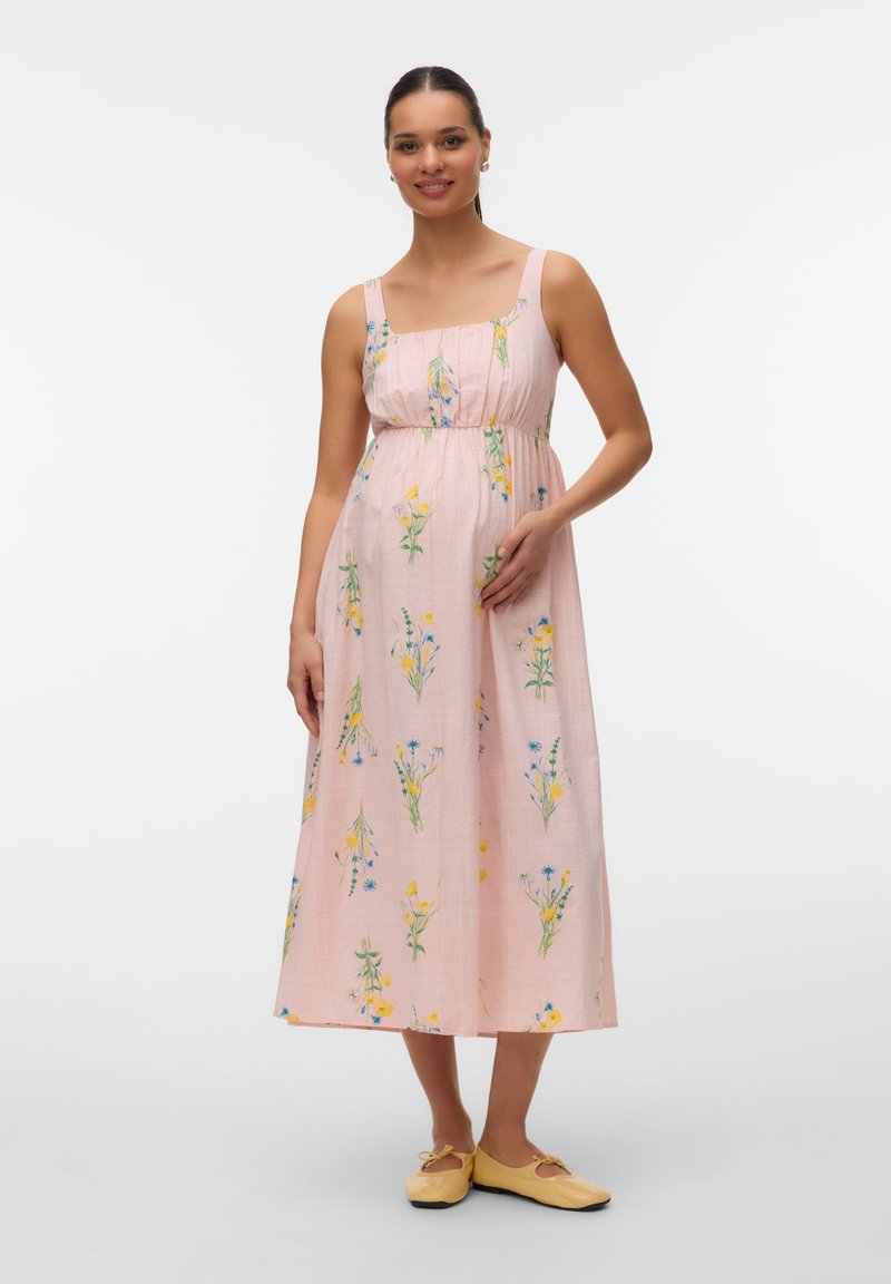 Χαμογελαστή γυναίκα σε ροζ φλοράλ αμάνικο μίντι φόρεμα και κίτρινα φλατ παπούτσια, όρθια με το ένα χέρι να ακουμπά στην κοιλιά της.