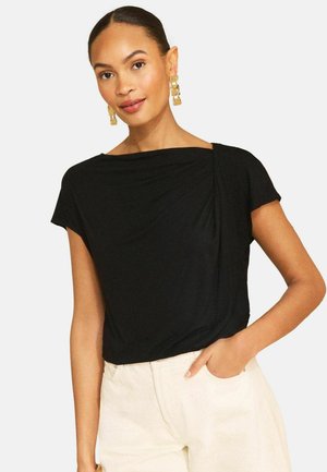 Vrouw draagt een zwarte top met korte mouwen en een gedrapeerde halslijn en gouden rechthoekige oorbellen, met één hand in de zak van een lichte broek.