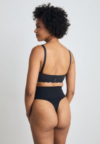 Svart blonde bralette med bølgete kanter og tynne stropper, kombinert med høyhals svart truse. Glatt tekstur og tettsittende design.