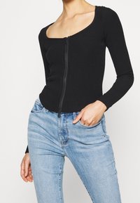 Sort ribbet langærmet top med firkantet halsudskæring og lynlåsdetalje foran, båret med lyseblå jeans. Har en tætsiddende design.