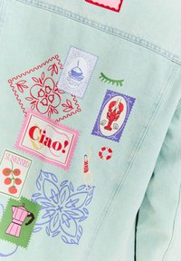 Jasnoniebieska kurtka dżinsowa z kolorowymi, dekoracyjnymi naszywkami przedstawiającymi różne wzory, w tym motywy jedzenia, natury i wakacji.