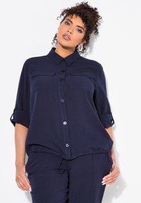 Camisa de botones azul marino con mangas recogibles, dos bolsillos en el pecho, cintura elástica y textura de tela suave. Presenta botones contrastantes.