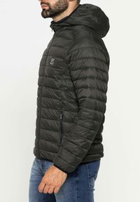 Giacca a vento nera imbottita con cappuccio, dotata di sezioni trapuntate, zipper frontale e un piccolo logo sul petto. Materiale leggero e testurizzato.