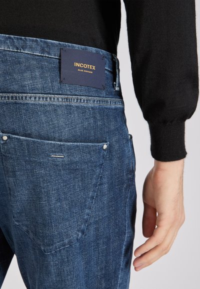 Jeansy o ciemnoniebieskim praniu, z doszytą metką na pasie oraz dwoma tylnymi kieszeniami z widocznymi nitami.