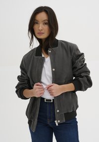 Gray leather bomber jacket with a front zipper, ribbed cuffs, and collar. Worn over a white t-shirt, paired with dark blue jeans.