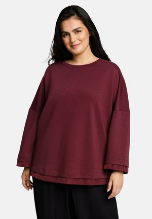 Woman with long dark hair wearing a loose, long-sleeve burgundy top and black pants, smiling with hands relaxed by sides.