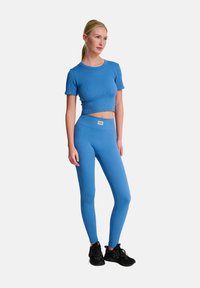 Niebieski prążkowany top na krótko z wysokim stanem, w połączeniu z niebieskimi legginsami o wysokim stanie. Oba mają gładką teksturę i małą metkę z logo na pasie.