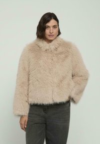 Kobieta z krótkimi, ciemnymi włosami, ubraną w beżową, puszystą kurtkę i ciemnoszare spodnie, stojąca na tle jednolitego, jasnego tła.